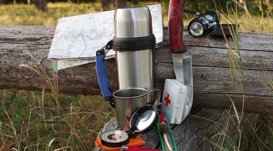 A group of camping items on a log, a map, compass, first aid kit, flashlight,  thermos. 